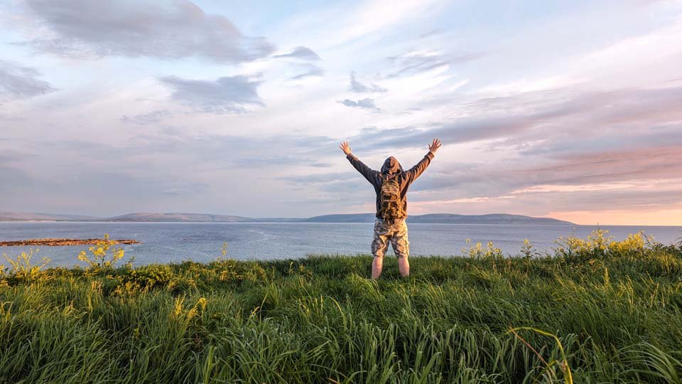 Male hiker in camo shorts and backpack on top of green hill, hands up, view on wild Atlantic way at Galway, Ireland